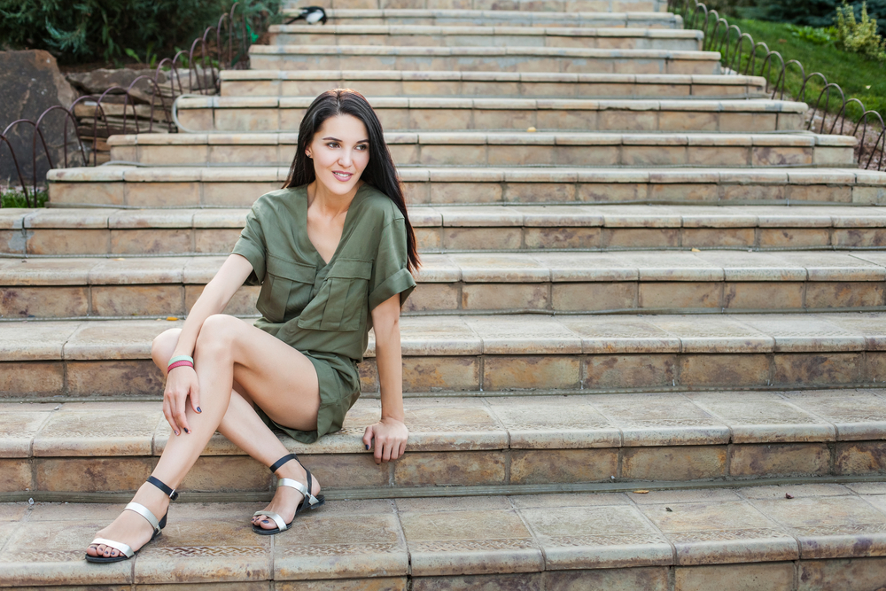 A young woman sits on a stairs in a green romper and black and silver sandals. 