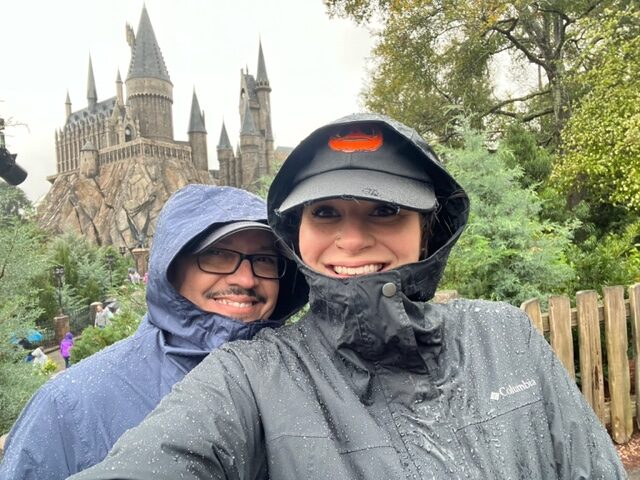 A couple poses in the rain at Universal Studios: their ponchos are keeping them dry as they smile with Hogwarts in the background. 