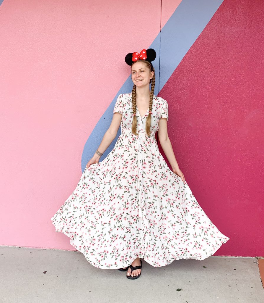 In front of the pink wall at Disney, a woman in Minnie ears and a maxi floral dress smiles, comfortable that she chose to wear this when considering what to wear in Orlando. 