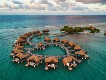 overwater bungalows with thatched roofs in a heart shape. it is sunset out so the sky is cloudy but the colors are pretty, reminiscent of the Caribbean