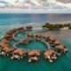 overwater bungalows with thatched roofs in a heart shape. it is sunset out so the sky is cloudy but the colors are pretty, reminiscent of the Caribbean