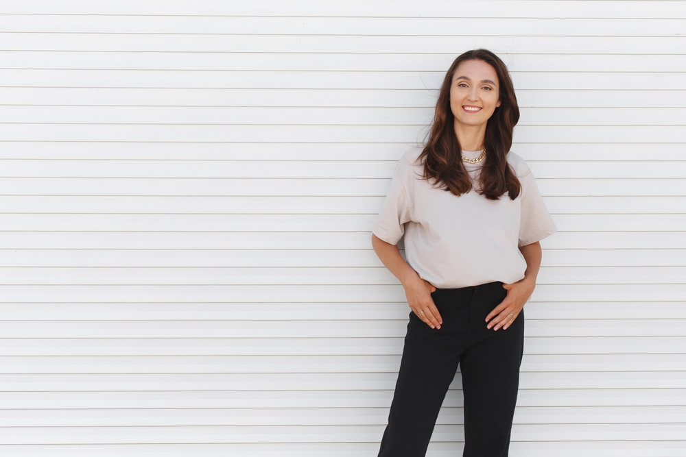 A woman poses with her hands in her pocket, enjoying the weather and her beige shirt, as well as her casual black pants: she has added a statement necklace for an extra flare. 