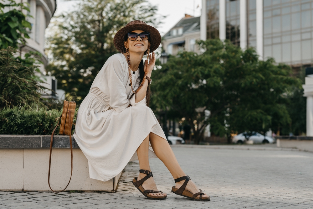 A woman in a linen, white midi dress sits on the edge of a sidewalk with her crossbody bag, sandals, and woven hat. 