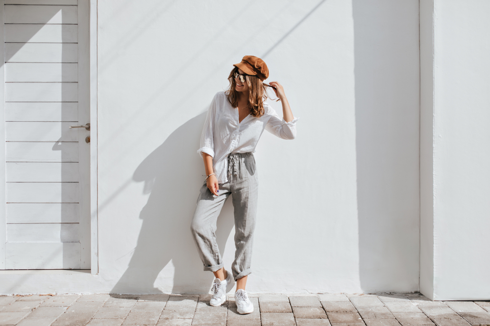 A woman in linen pants, white sneakers, and a button down white shirt smiles off camera while twirling her hair. Her brown cap is perfect for what to wear in Tampa. 