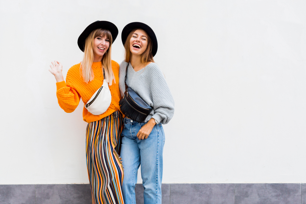 Two friends pose against a white back drop: one wears jeans and a grey sweater, while another wears a striped skirt and an orange sweater. Both wear a black hat and wear fanny packs across their chests. 