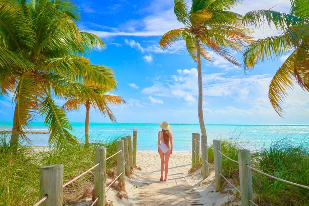 A woman in a one piece white suit stands on the cusp of the beach: the water is vibrantly blue beyond her.