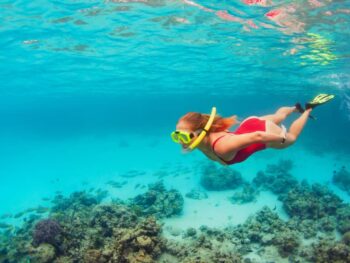 woman wearing a red bathing suit and a yellow mask and snorkel swimming under the water. the water is clear and blue!