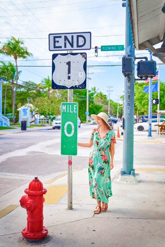 A girl in a floor length floral dress smiles next to Mile zero sign in key west, a popular destination stop, a place to visit but not spend too much time at, as per mistakes to avoid in Key West.