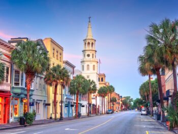 At dusk, the downtown area of Charleston is quaint and artsy, with fun colored buildings to explore. the street is lined with palm trees and there is a white church in the middle