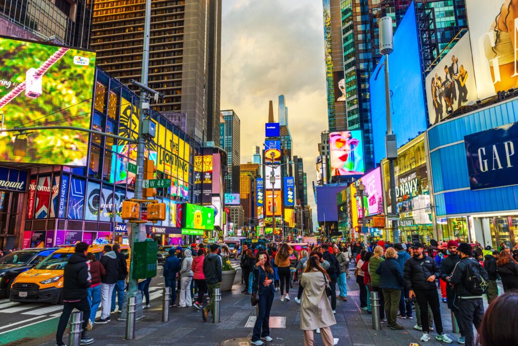 Tons of tourists gather in times square as the sun goes down, lights on billboards bright to match the coats of the tourists.