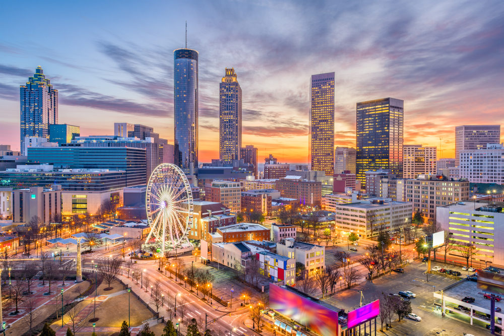 An overhead shot of the Atlanta skyline shows tall skyscrapers, a ferris wheel and more at dusk, the lights coming alive.