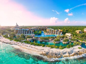 An overhead shot of Cancun shows the crystal clear water lapping against the shore as tall resorts tower beyond the coast, the water is turquoise and the sky is blue
