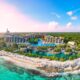 An overhead shot of Cancun shows the crystal clear water lapping against the shore as tall resorts tower beyond the coast, the water is turquoise and the sky is blue