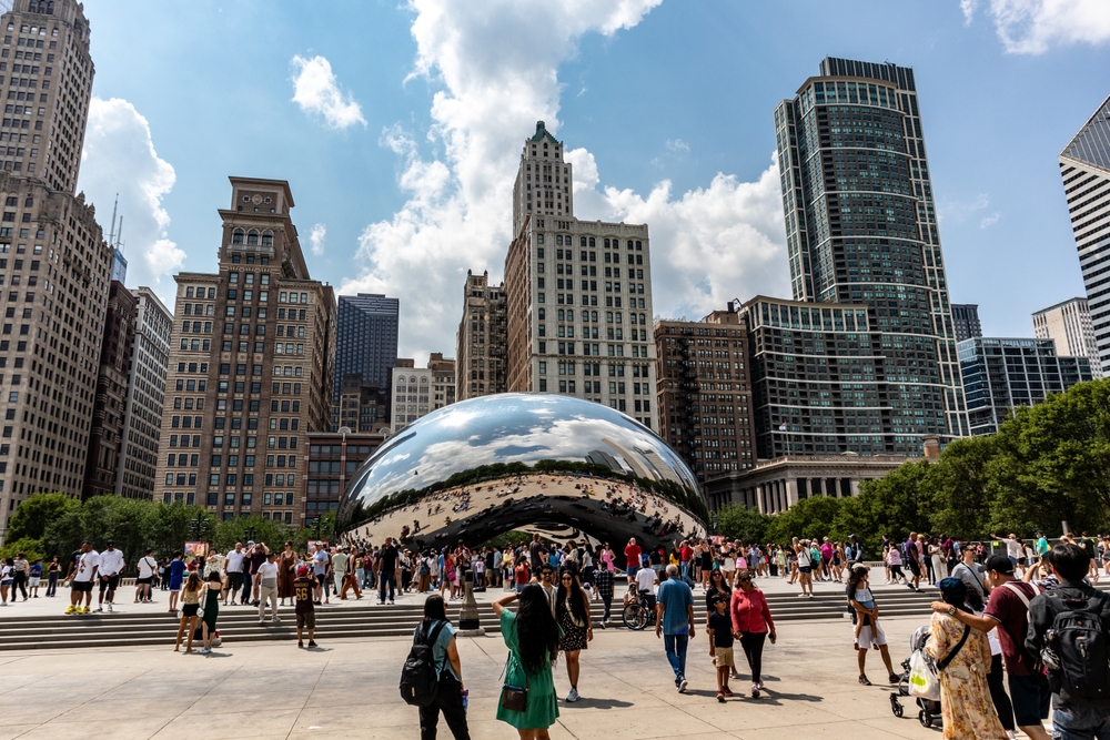 Crowds gather around the iconic Chicago bean, taking photos, smiling, skyscrapers all around them. Chicago is one of the cheapest places to fly from Orlando.