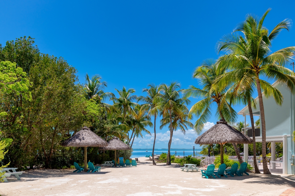 At Key West, tiki huts line concrete areas that then lead into sandy beaches with clear blue water. Palm trees stand tall and green.