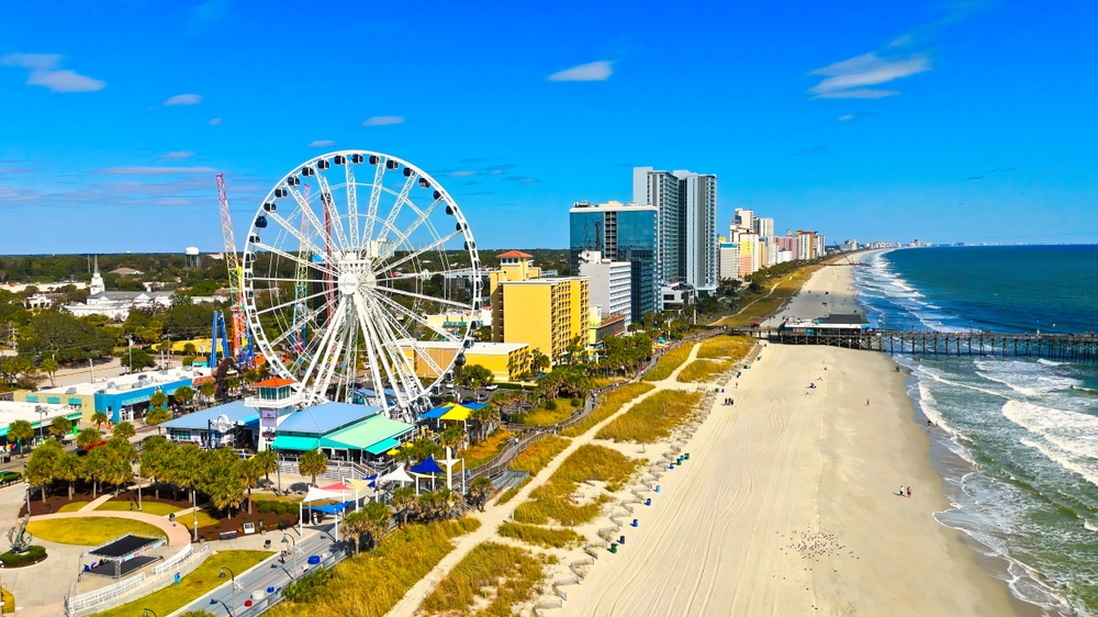 An overhead shot of Myrtle beach shows the coast with a boardwalk, hotels, blue skies, sandy beaches, and ferris wheel.