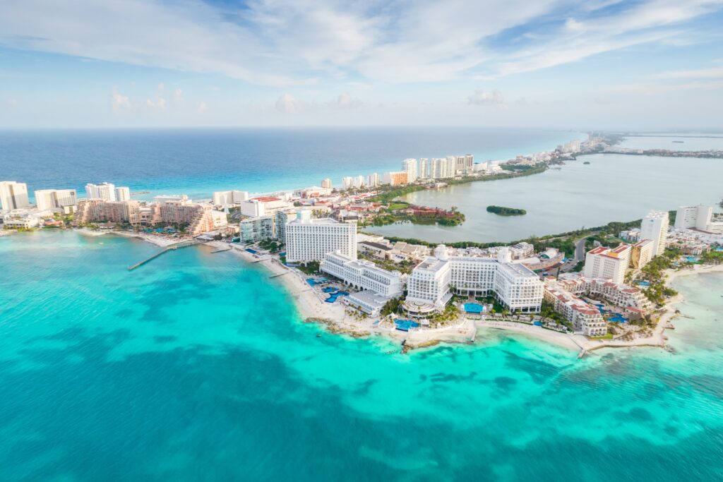 An overhead shot of Cancun, Mexico shows a strip of the beach lined with hotels and pools and beaches with clear blue water. 