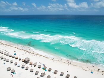 Vacation riviera maya cancun beach. The article is about cheapest places to fly from Florida. there is a beach with beautiful blue water and lots of brown umbrellas made out of tiki material along the beach. people are on the beach and in the water and the sky is blue with white clouds
