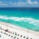 Vacation riviera maya cancun beach. The article is about cheapest places to fly from Florida. there is a beach with beautiful blue water and lots of brown umbrellas made out of tiki material along the beach. people are on the beach and in the water and the sky is blue with white clouds