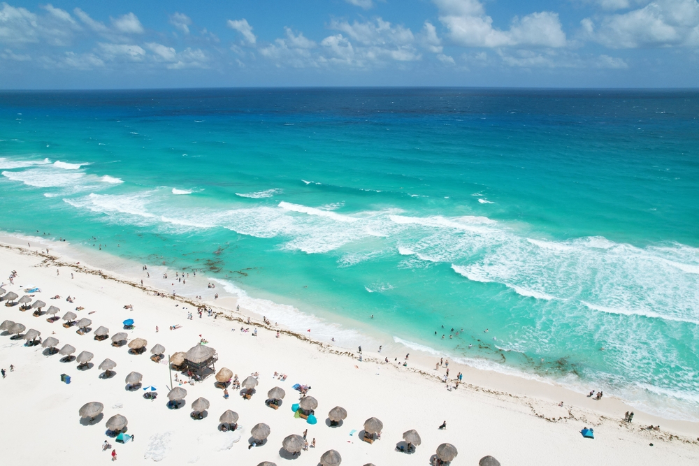 Vacation riviera maya cancun beach. The article is about cheapest places to fly from Florida. there is a beach with beautiful blue water and lots of brown umbrellas made out of tiki material along the beach. people are on the beach and in the water and the sky is blue with white clouds