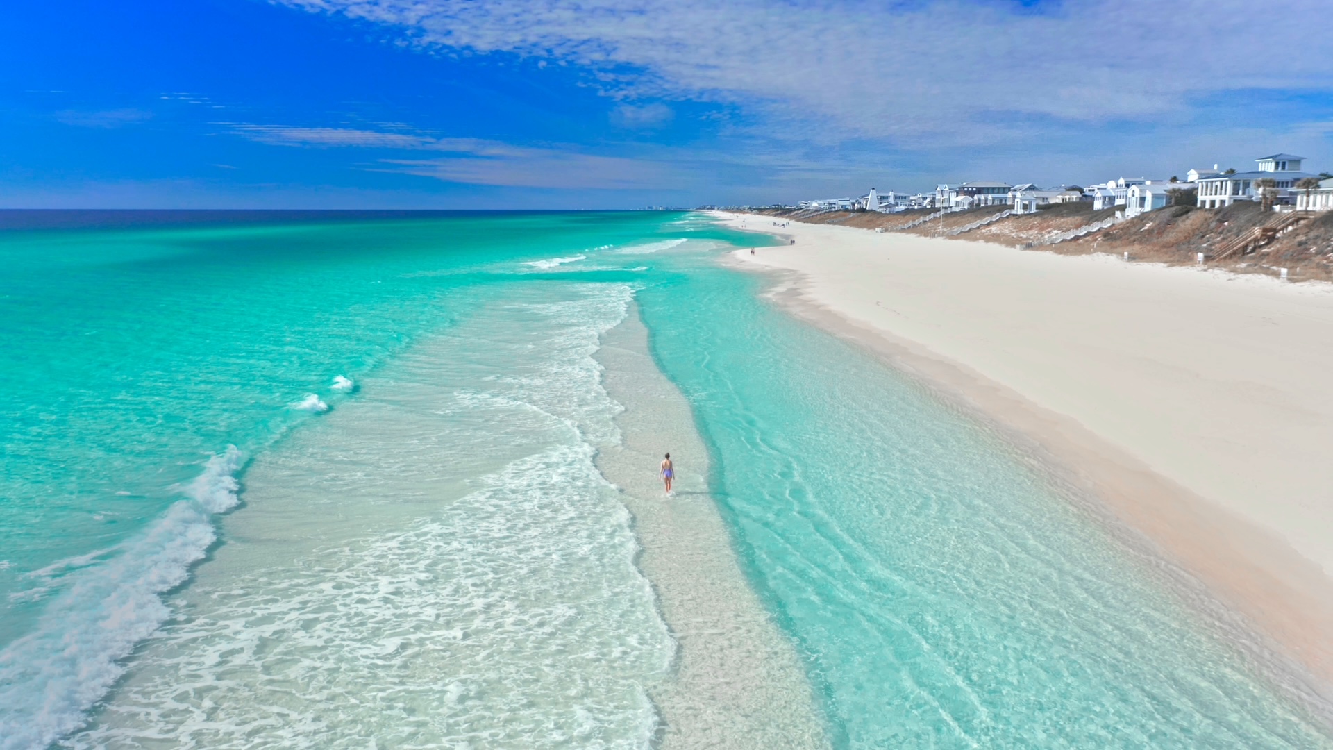 Aerial view of 30A beach with emerald green water and gentle waves rolling onto wide white sand. A single person walks along the shallow shoreline with coastal homes lining the dunes under a partly cloudy blue sky.
